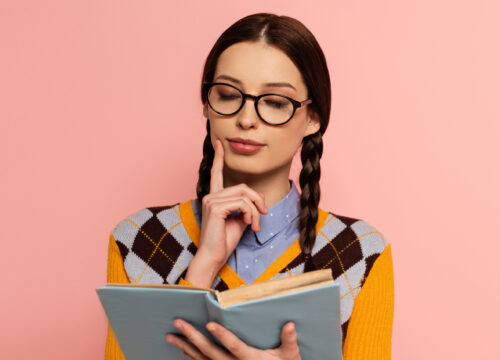 Woman with glasses and braids reading a book on a pink background.